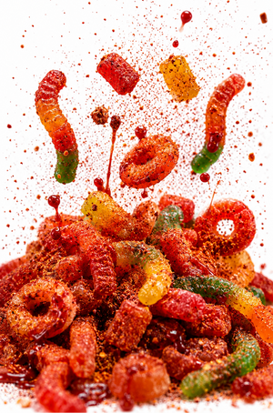 A pile of colorful gummy worms and ring-shaped gummies coated in red chili powder, with pieces splashing into the air against a white background.